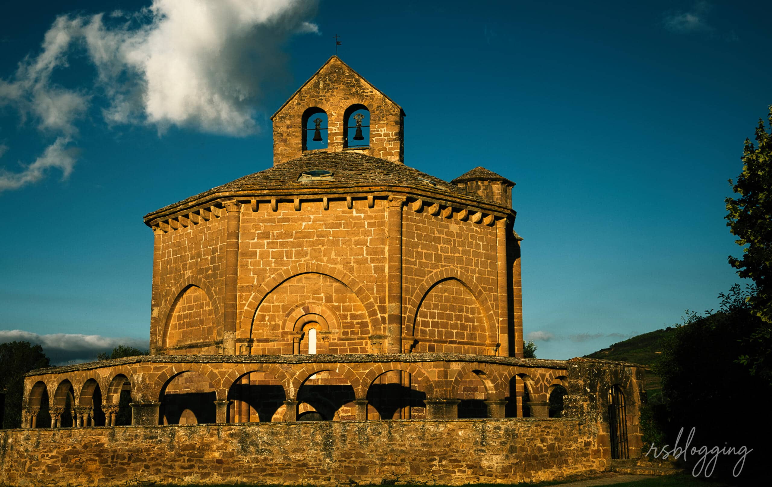 Ermita de Santa María de Eunate iluminada por el sol de la tarde, con cielo azul intenso y nubes al fondo